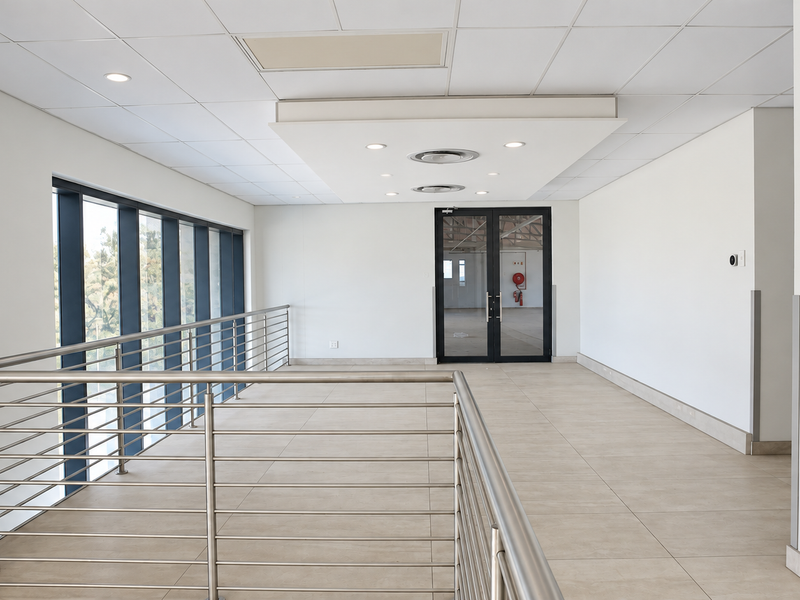 Modern office building hallway featuring tiled flooring, stainless steel balustrades, recessed ceiling lighting, and access to an elevator and double glass doors.