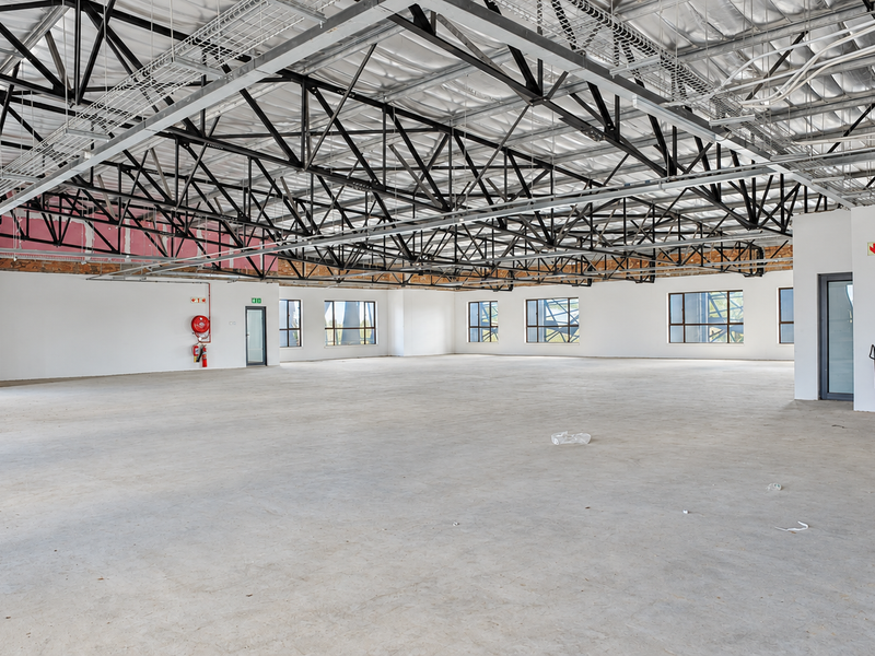 A large, open-plan office space featuring industrial-style exposed steel roof trusses, concrete flooring, and ample natural light through multiple windows.