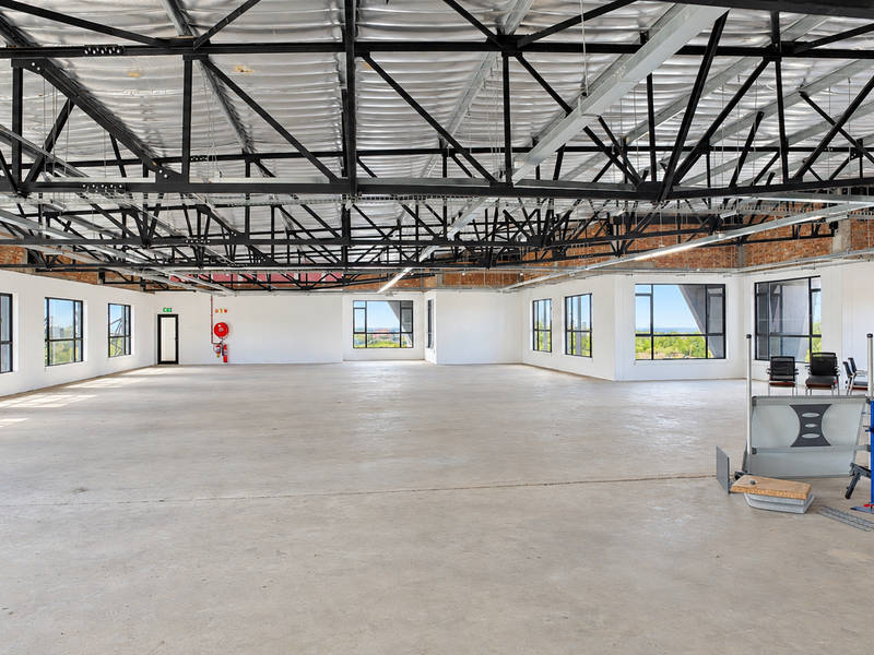 A spacious, open-plan office area featuring high ceilings with exposed steel trusses, polished concrete floors, and large windows providing ample natural light.