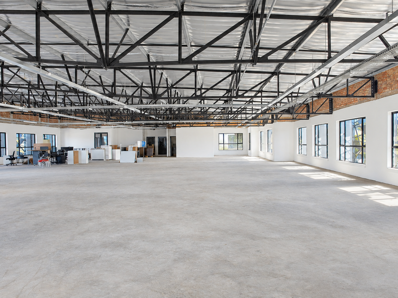 Spacious open-plan office area featuring high exposed steel roof trusses, polished concrete floors, and abundant natural light through large windows.