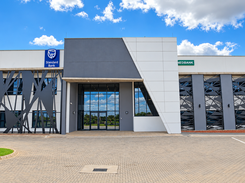 Modern commercial office building facade featuring contemporary architectural design, glass entrance, and decorative metal screening with paved parking area.