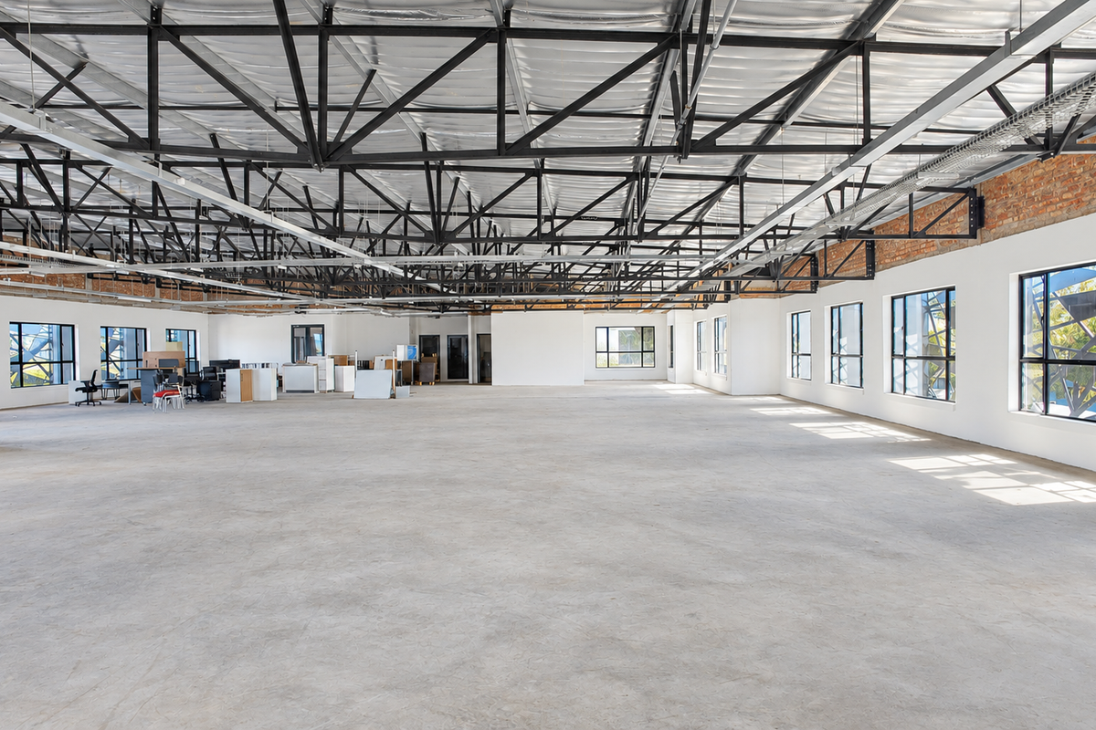 Spacious open-plan office area featuring high exposed steel roof trusses, polished concrete floors, and abundant natural light through large windows.