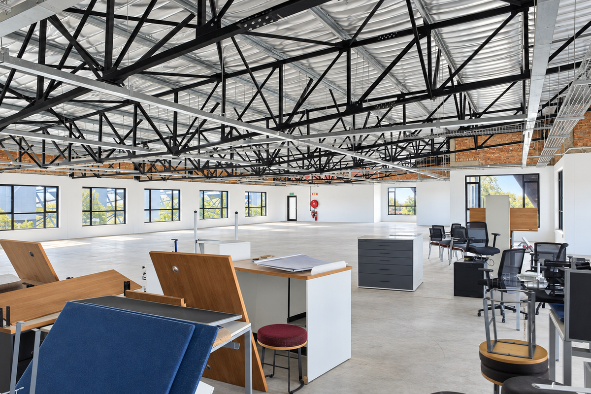 Spacious open-plan office featuring high exposed steel roof trusses, polished concrete flooring, and large windows providing ample natural light.