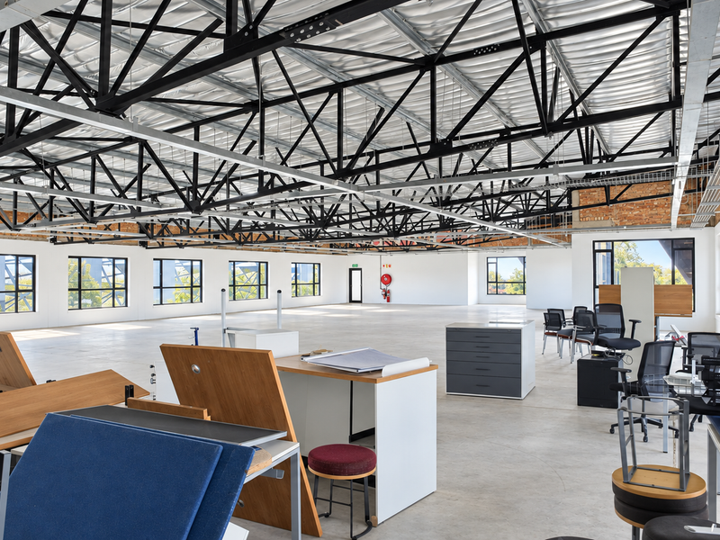 Spacious open-plan office featuring high exposed steel roof trusses, polished concrete flooring, and large windows providing ample natural light.