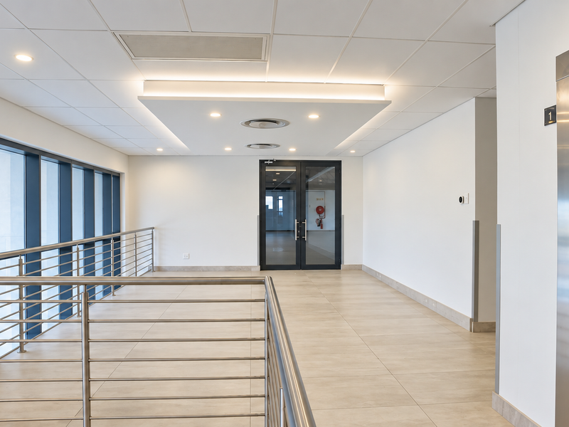 A modern, well-maintained office hallway featuring tiled flooring, a suspended ceiling with recessed lighting, a stainless steel balustrade, and access to an elevator.