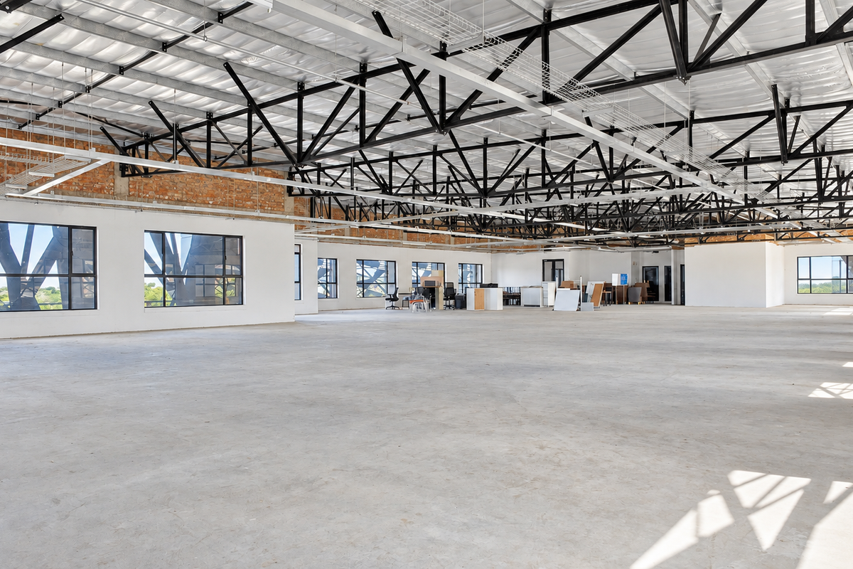 A spacious, open-plan office area featuring high ceilings with exposed steel trusses, polished concrete floors, and large windows providing ample natural light.