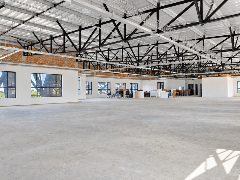 A spacious, open-plan office area featuring high ceilings with exposed steel trusses, polished concrete floors, and large windows providing ample natural light.