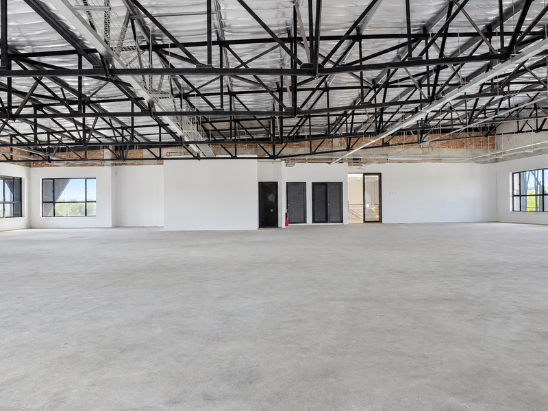 Spacious open-plan office area featuring high exposed steel roof trusses, polished concrete floors, and large windows providing ample natural light.