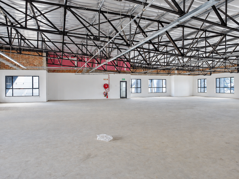 A large, open-plan office space featuring high exposed steel roof trusses, concrete flooring, and ample natural light through multiple aluminium-framed windows.