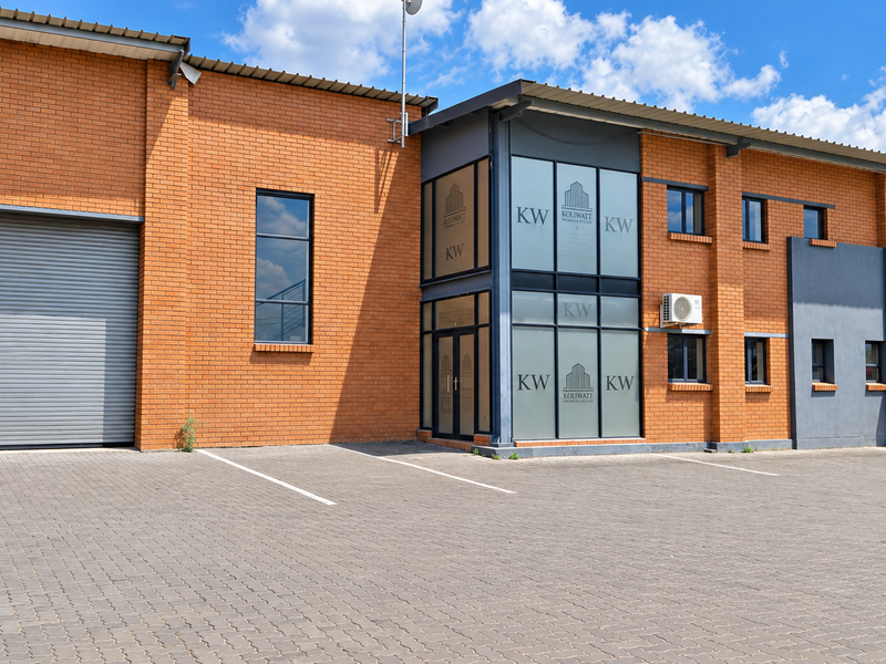 Modern industrial property featuring a brick facade, large roller shutter door, and a prominent glass-fronted entrance with paved parking area.