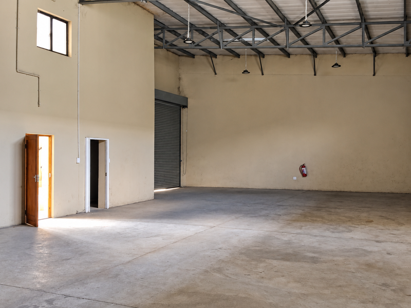 Spacious industrial warehouse unit featuring high ceilings with exposed steel trusses, concrete flooring, and a large roller shutter door for easy access.