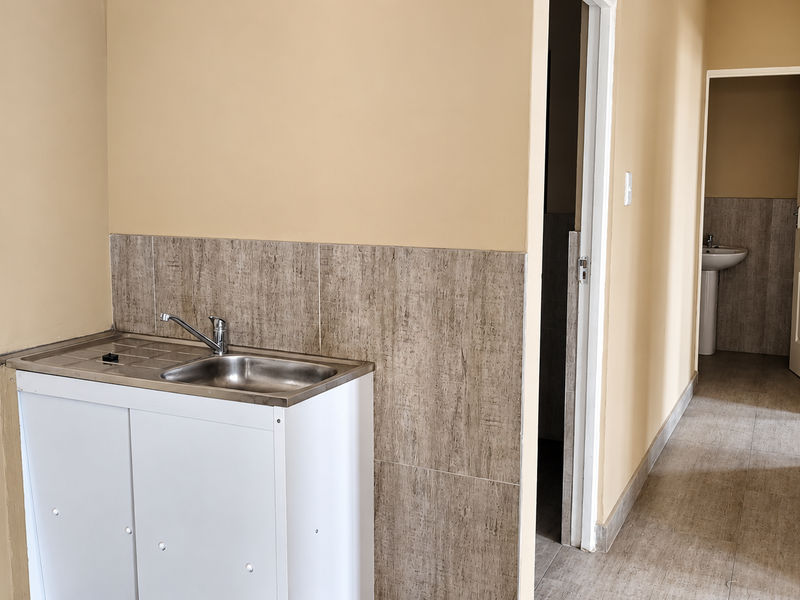 A functional kitchenette area featuring a stainless steel sink with tiled splashback, leading into a hallway with access to a bathroom with pedestal basin.
