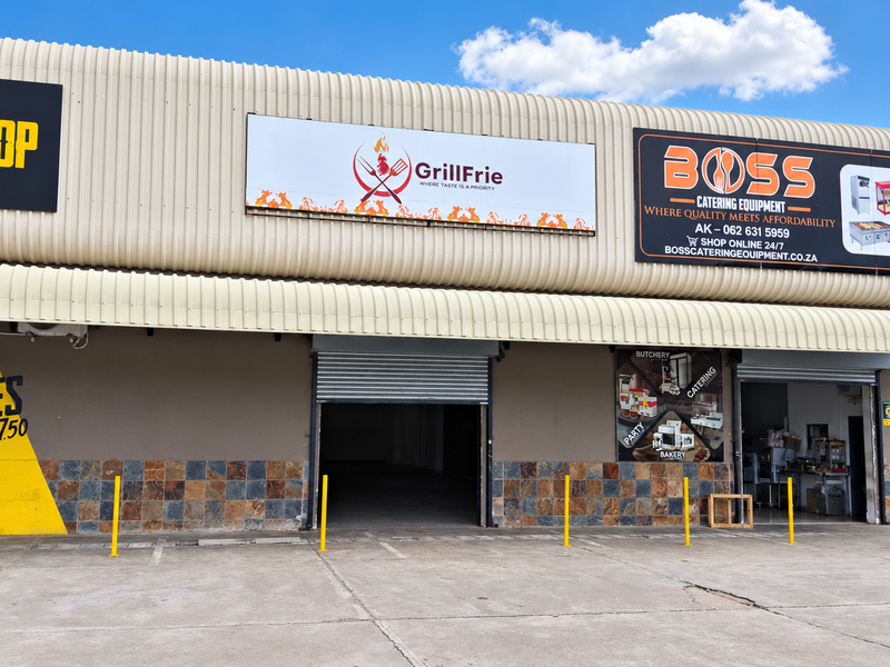 Front elevation of a retail unit featuring corrugated metal cladding, tiled lower walls, and a roller shutter entrance within a commercial complex.