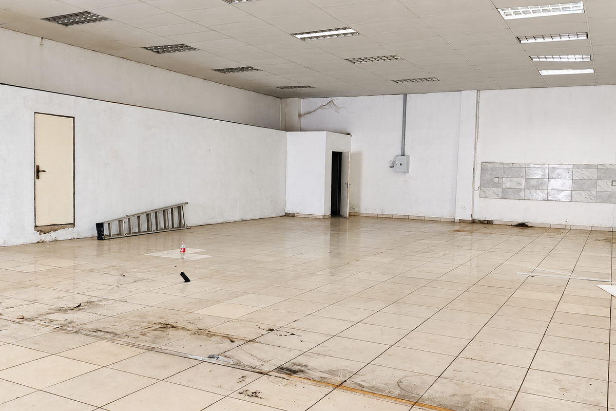 A large, vacant open-plan retail space featuring tiled flooring, a suspended ceiling with recessed lighting, and a dedicated tiled splashback area.