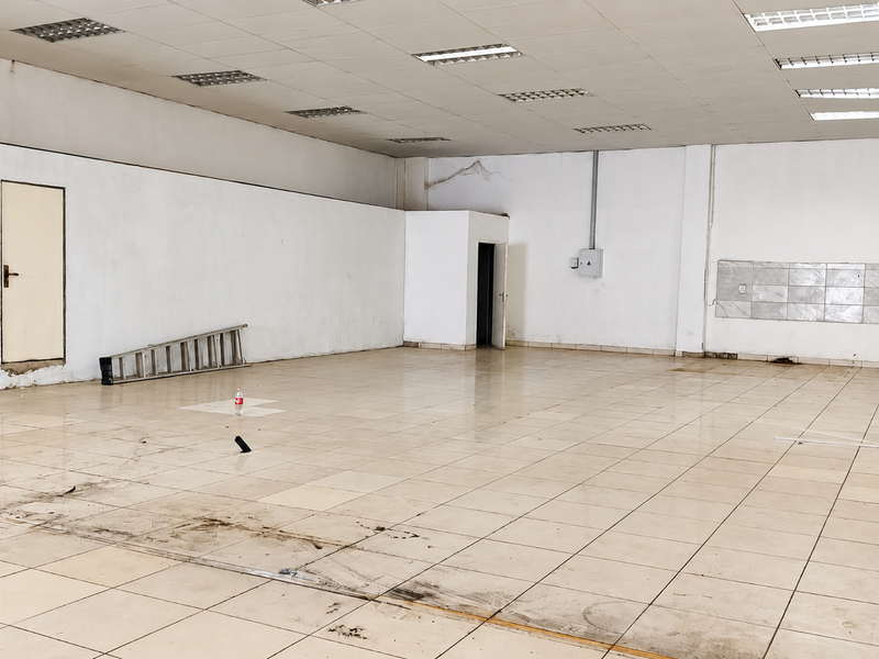 A large, vacant open-plan retail space featuring tiled flooring, a suspended ceiling with recessed lighting, and a dedicated tiled splashback area.