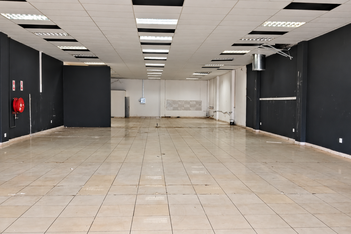 A spacious, empty retail unit featuring tiled flooring, a suspended ceiling with recessed lighting, and a wall-mounted fire hose reel.