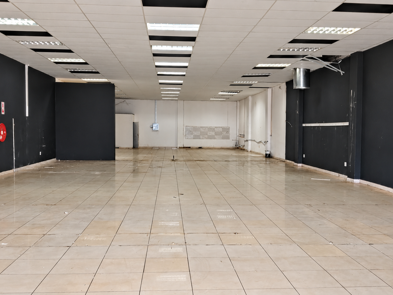 A spacious, empty retail unit featuring tiled flooring, a suspended ceiling with recessed lighting, and a wall-mounted fire hose reel.