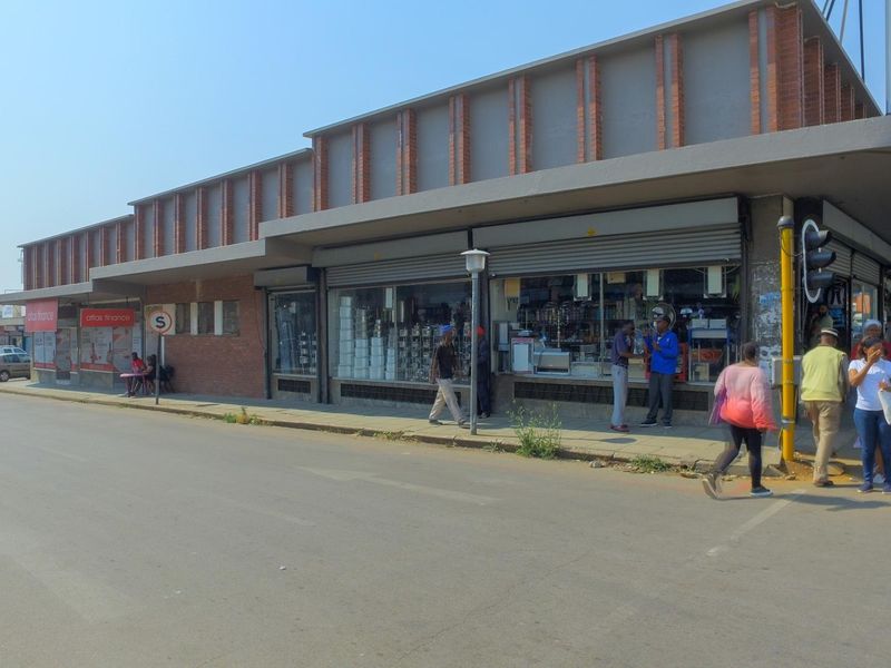 Street-level view of a commercial retail building featuring brick facade, large display windows, and covered pedestrian walkway in a busy urban setting.