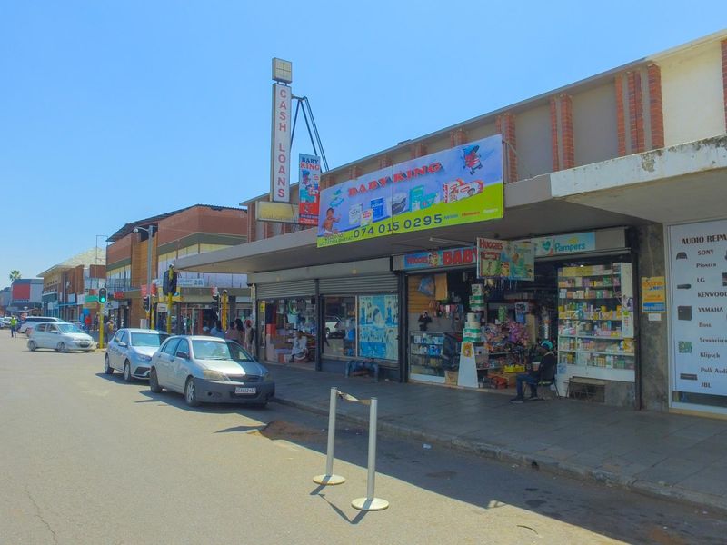 Street-level view of a commercial retail building featuring multiple shopfronts with signage, display windows, and a covered pedestrian walkway.