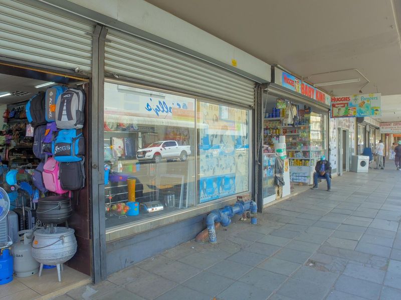 Street-level view of a commercial retail shopfront featuring large display windows, roller shutter doors, and a paved pedestrian walkway.