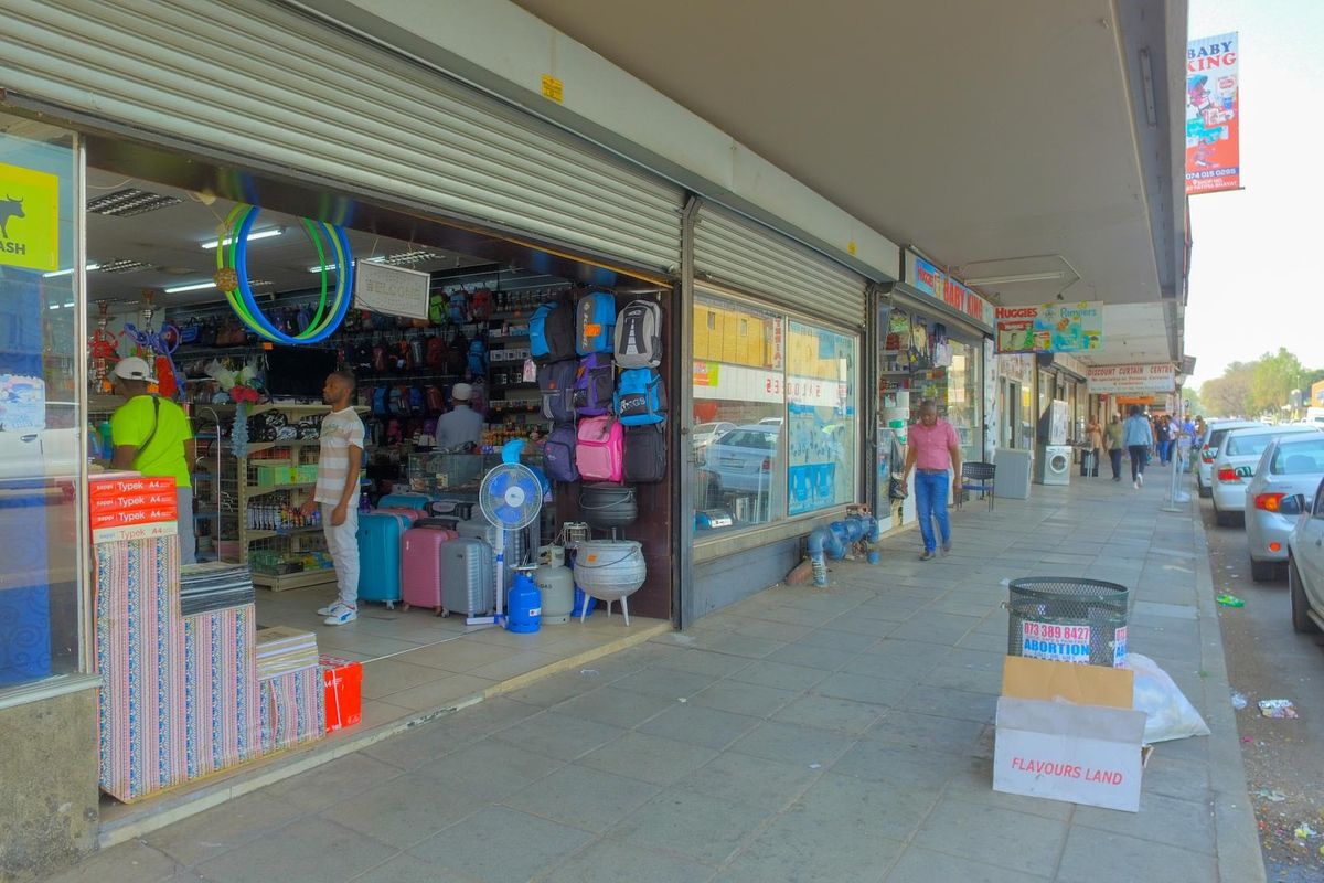 Street-level view of a commercial storefront featuring large display windows and metal roller shutter doors along a paved pedestrian walkway.