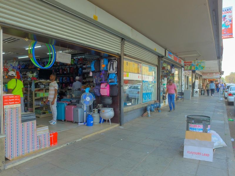 Street-level view of a commercial storefront featuring large display windows and metal roller shutter doors along a paved pedestrian walkway.