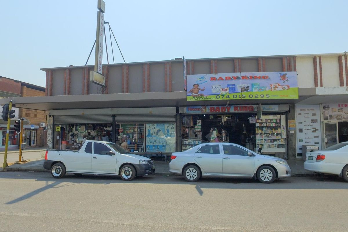 Street-level view of a commercial retail shopfront featuring roller shutter doors, signage, and a covered walkway in a busy urban business district.