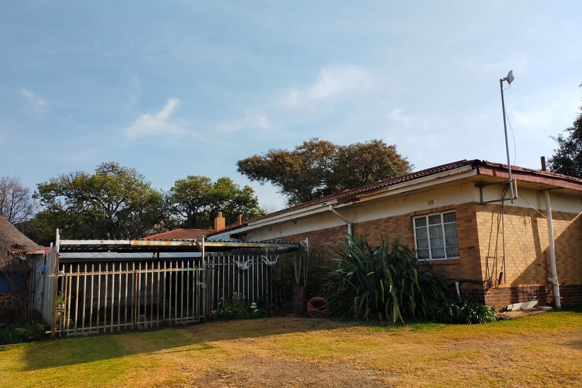 Rear view of a brick farmhouse featuring a covered carport area, metal security fencing, and established garden surroundings.