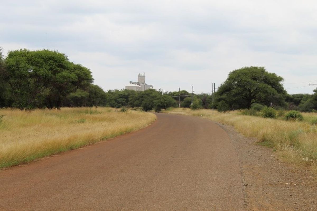 A view of a paved road leading through a rural landscape with surrounding vegetation and distant industrial infrastructure.