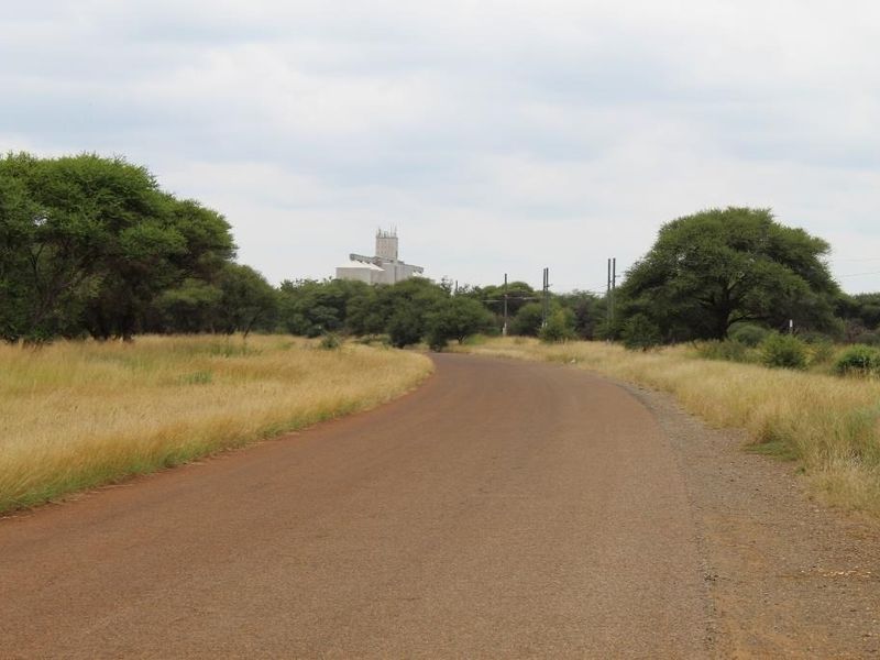 A view of a paved road leading through a rural landscape with surrounding vegetation and distant industrial infrastructure.
