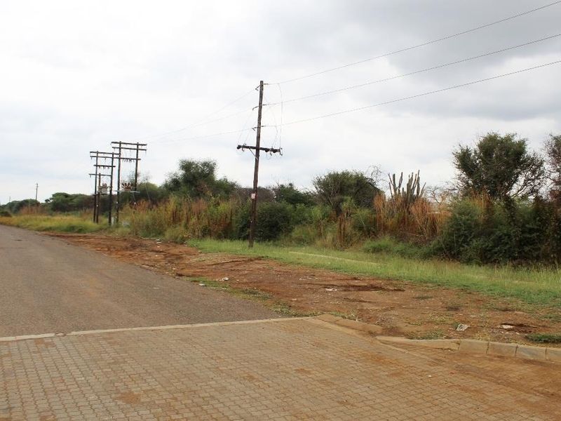 A street-level view of a vacant commercial plot featuring existing utility infrastructure, including power lines and poles, adjacent to a paved road.