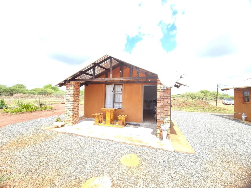 A rustic farm-style cottage featuring a covered patio with brick pillars, exposed timber rafters, and a corrugated metal roof set in a gravel yard.
