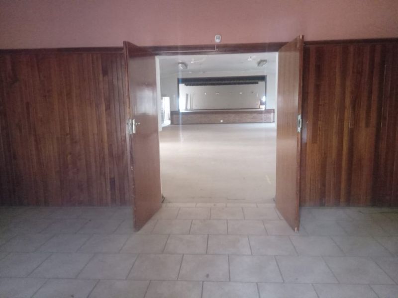 A spacious, empty commercial hall featuring wooden wall paneling, tiled floors, and a raised stage area, suitable for events or community gatherings.