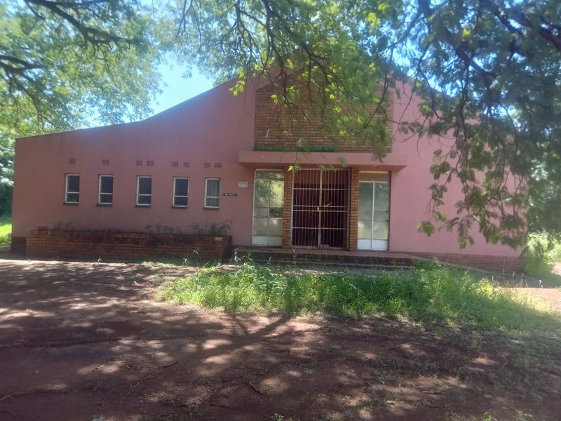 Front elevation of a commercial building featuring a brick facade, security gate entrance, and multiple windows, set within a natural, tree-lined environment.
