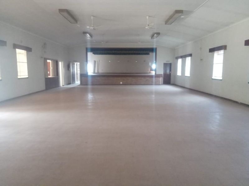 A large, vacant commercial hall featuring a raised stage, tiled flooring, and overhead lighting, suitable for events or community gatherings.