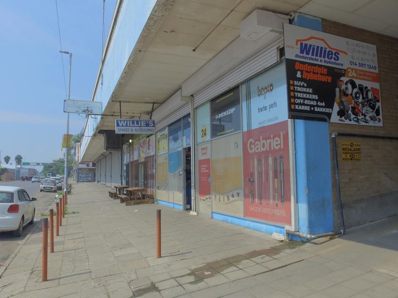Street-level view of a retail shopfront featuring large display windows, roller shutter security, and a covered walkway area.