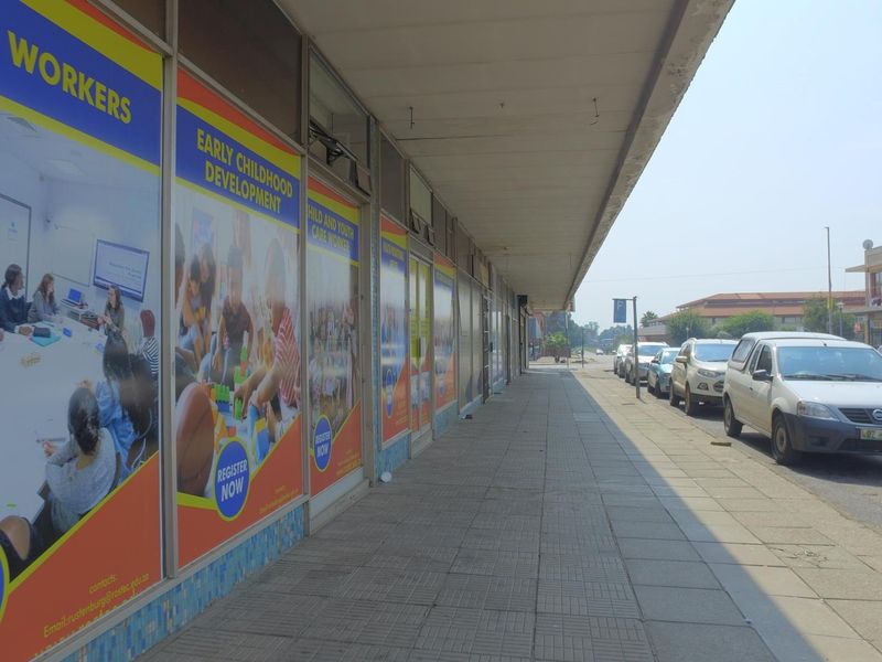 A street-level view of a retail storefront featuring a covered walkway, large display windows with promotional signage, and adjacent street parking.