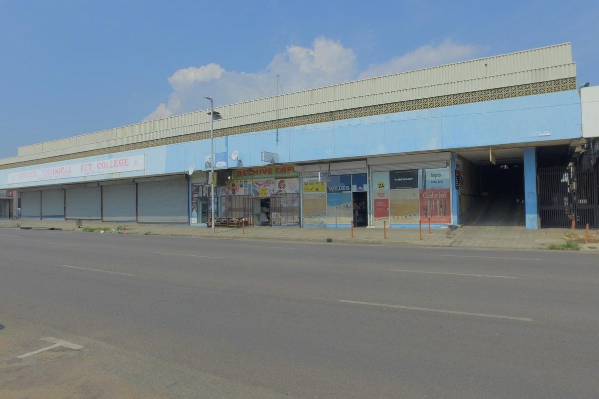 Street-level view of a commercial retail building featuring multiple storefronts with metal roller shutters and an integrated parking ramp access.