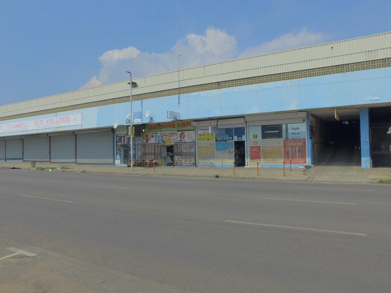 Street-level view of a commercial retail building featuring multiple storefronts with metal roller shutters and an integrated parking ramp access.
