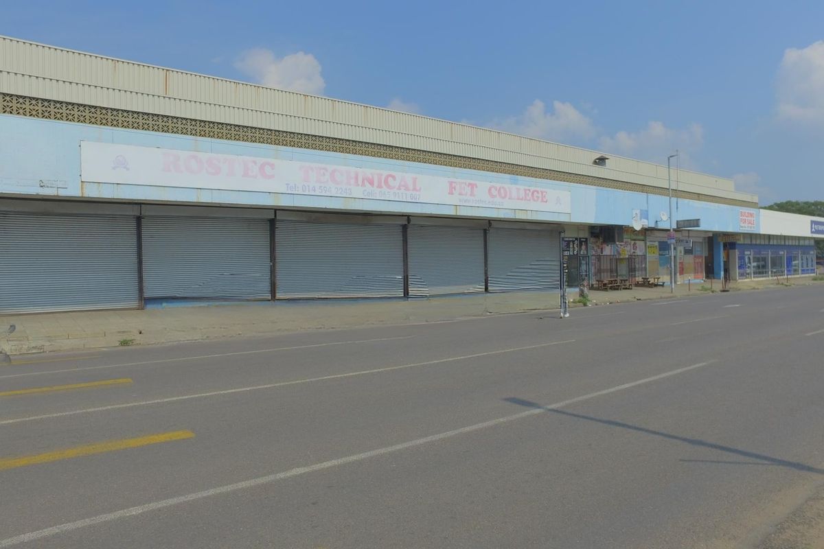 Street-level view of a commercial retail building featuring multiple metal roller shutter doors and prominent signage along the main road frontage.