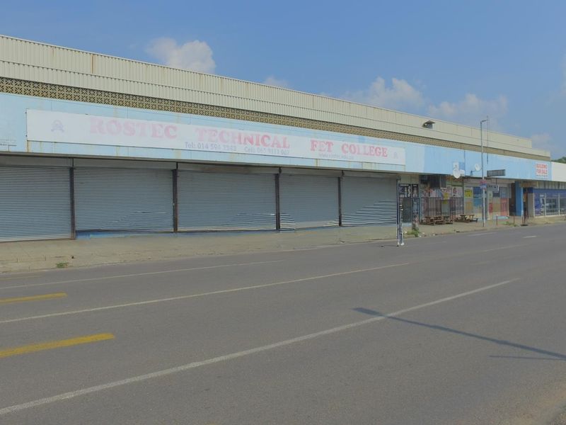 Street-level view of a commercial retail building featuring multiple metal roller shutter doors and prominent signage along the main road frontage.