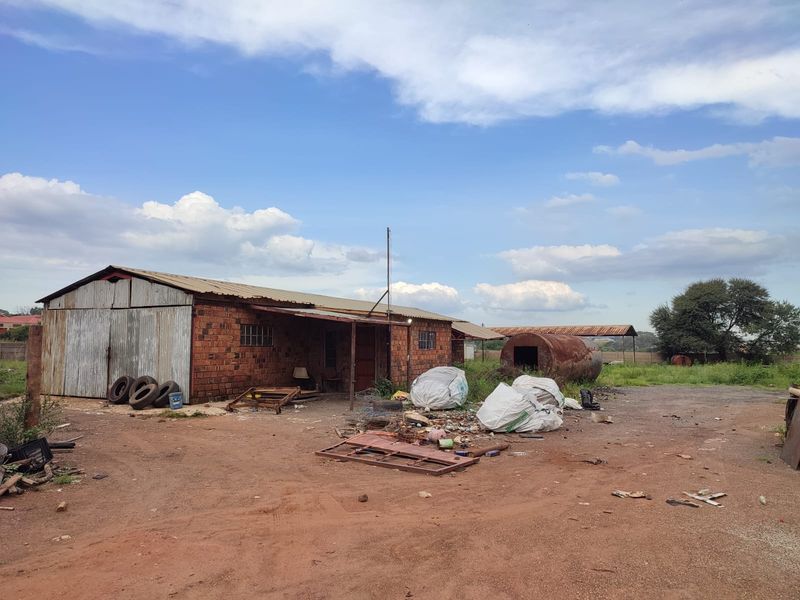 Industrial property featuring an exposed brick structure with corrugated metal roofing, sliding metal doors, and an adjacent rusted storage tank on a dirt lot.