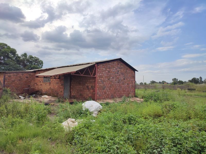 An industrial-style brick structure with a metal roof, situated on a large, overgrown plot of land.