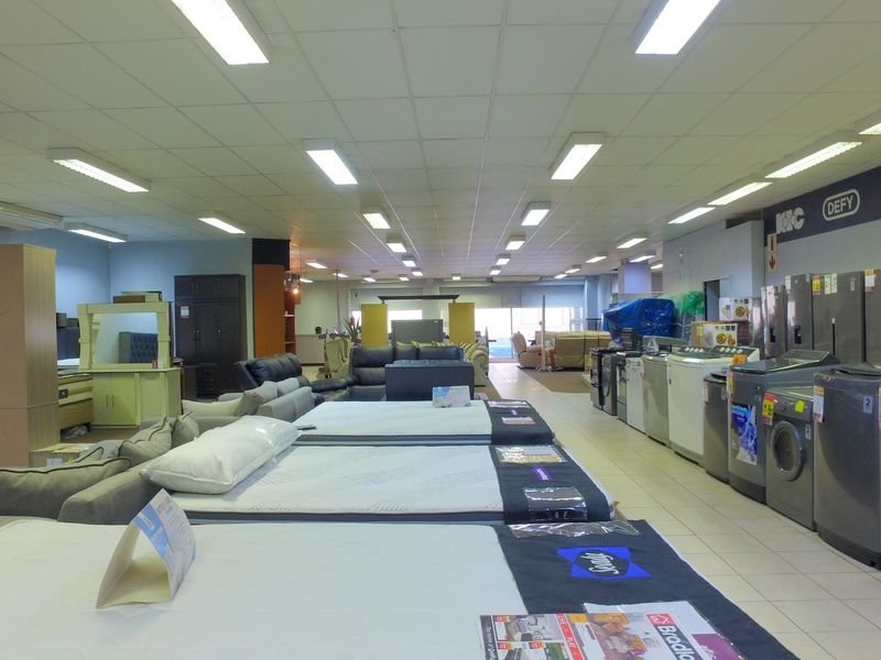 A spacious retail showroom floor featuring a suspended ceiling, fluorescent lighting, and tiled flooring, currently displaying various home appliances and furniture.