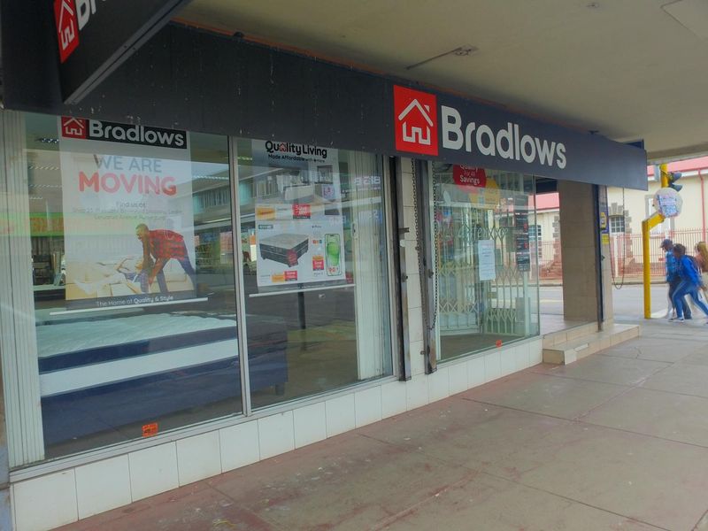 Street-level retail storefront featuring large display windows, prominent signage, and a covered walkway entrance in a commercial area.