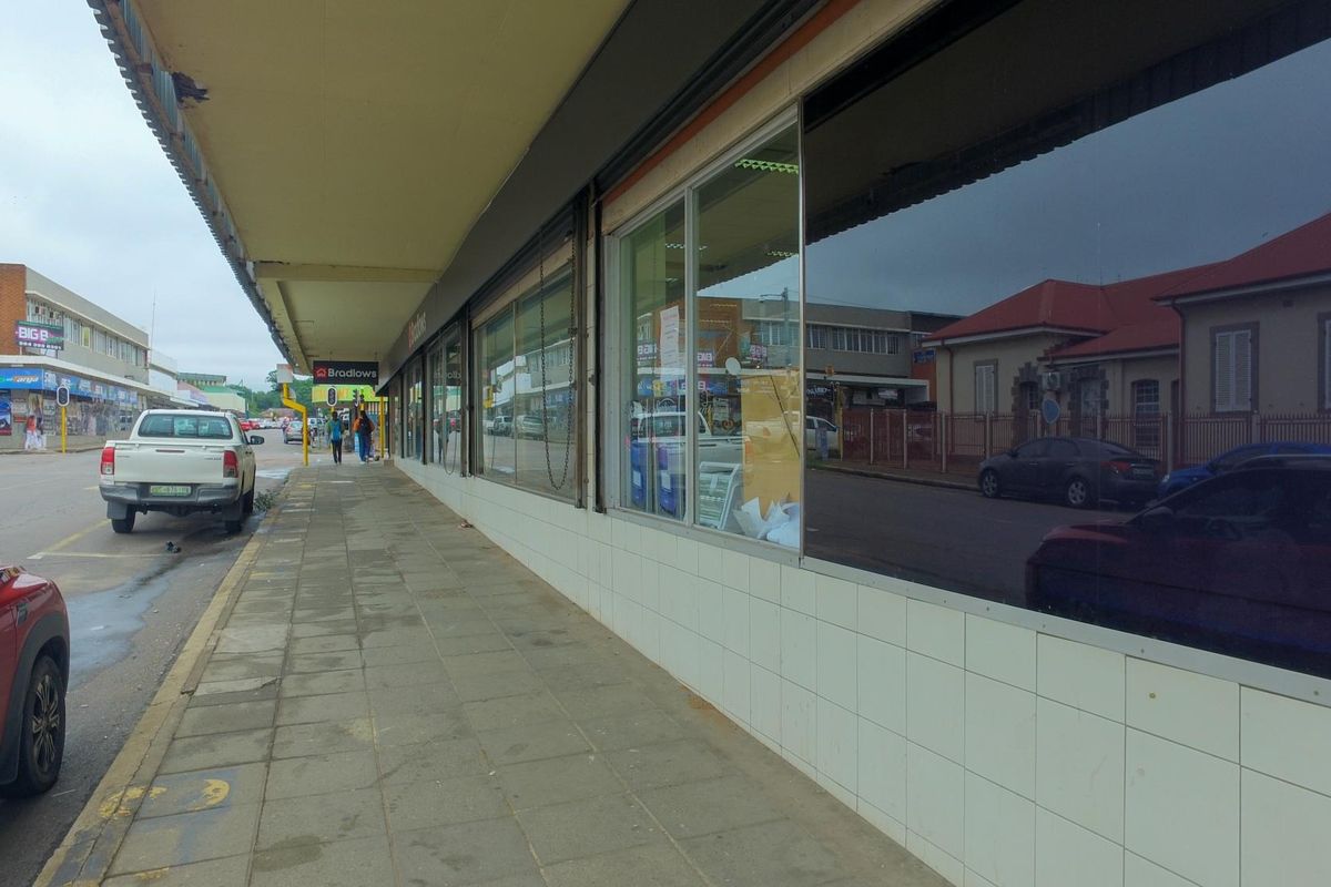 Street-level view of a retail storefront featuring large display windows, a covered walkway, and tiled facade base along a public sidewalk.