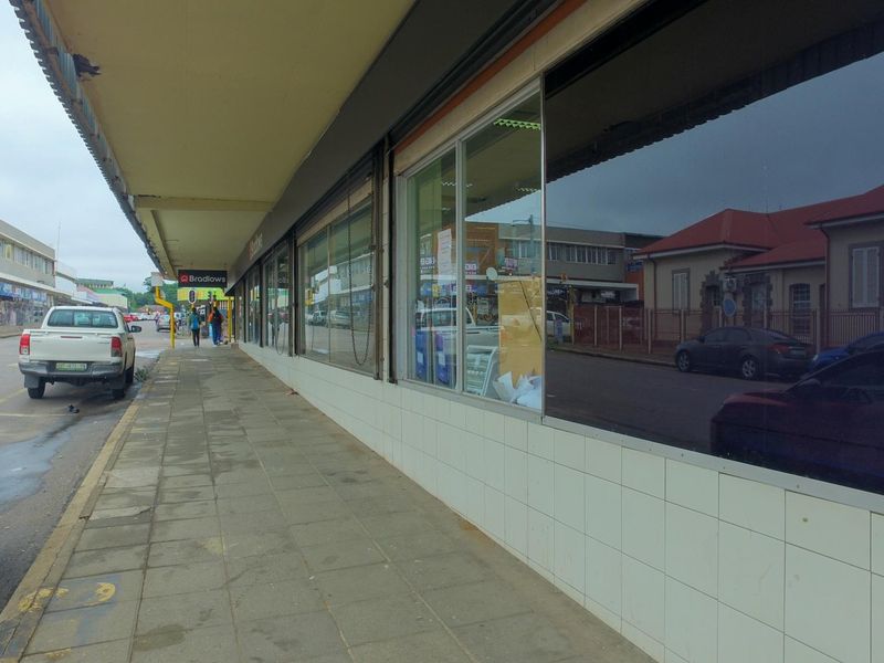Street-level view of a retail storefront featuring large display windows, a covered walkway, and tiled facade base along a public sidewalk.
