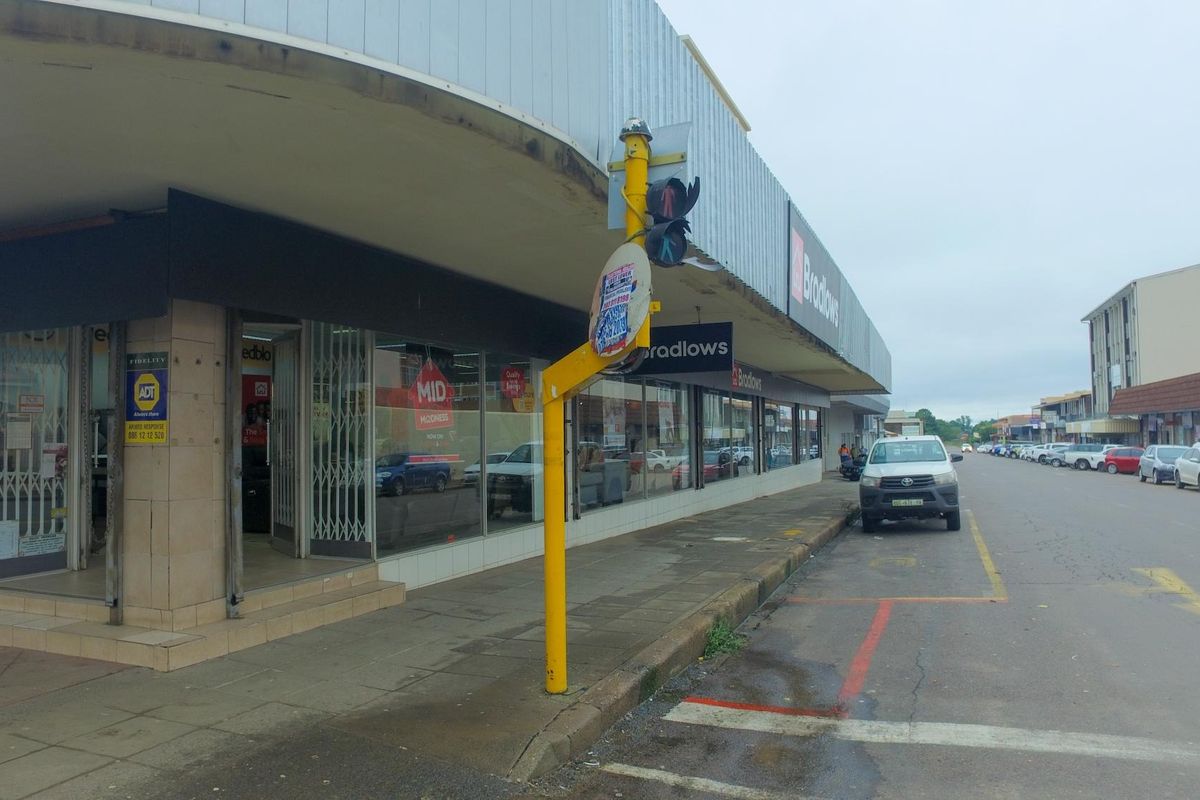 Street-level view of a retail storefront featuring large display windows, metal security gates, and a covered walkway in a commercial area.