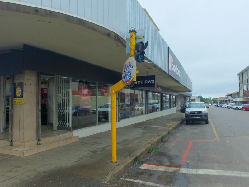 Street-level view of a retail storefront featuring large display windows, metal security gates, and a covered walkway in a commercial area.