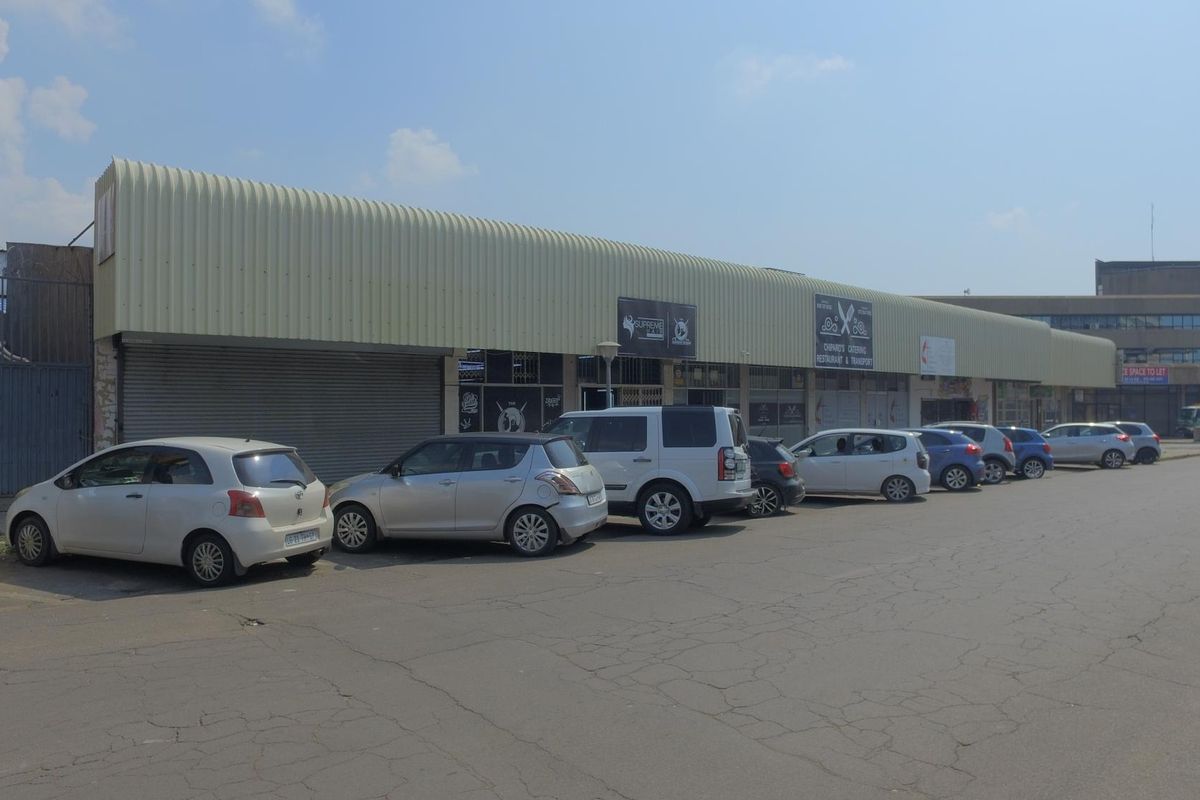 Front exterior view of a commercial retail strip center featuring a corrugated metal roof, roller shutter doors, and an adjacent paved parking area.